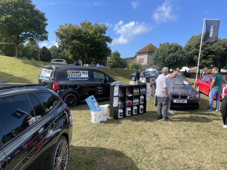 Valet Pro car care products display stand at an outdoor BMW car meet, with branded black van, detailing bottles and chemical jugs on shelf unit, alongside a dark purple BMW E92 and red classic car